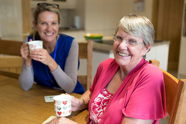 Community care worker joins an an elderly woman for a cup of tea while sitting at her kitchen table.